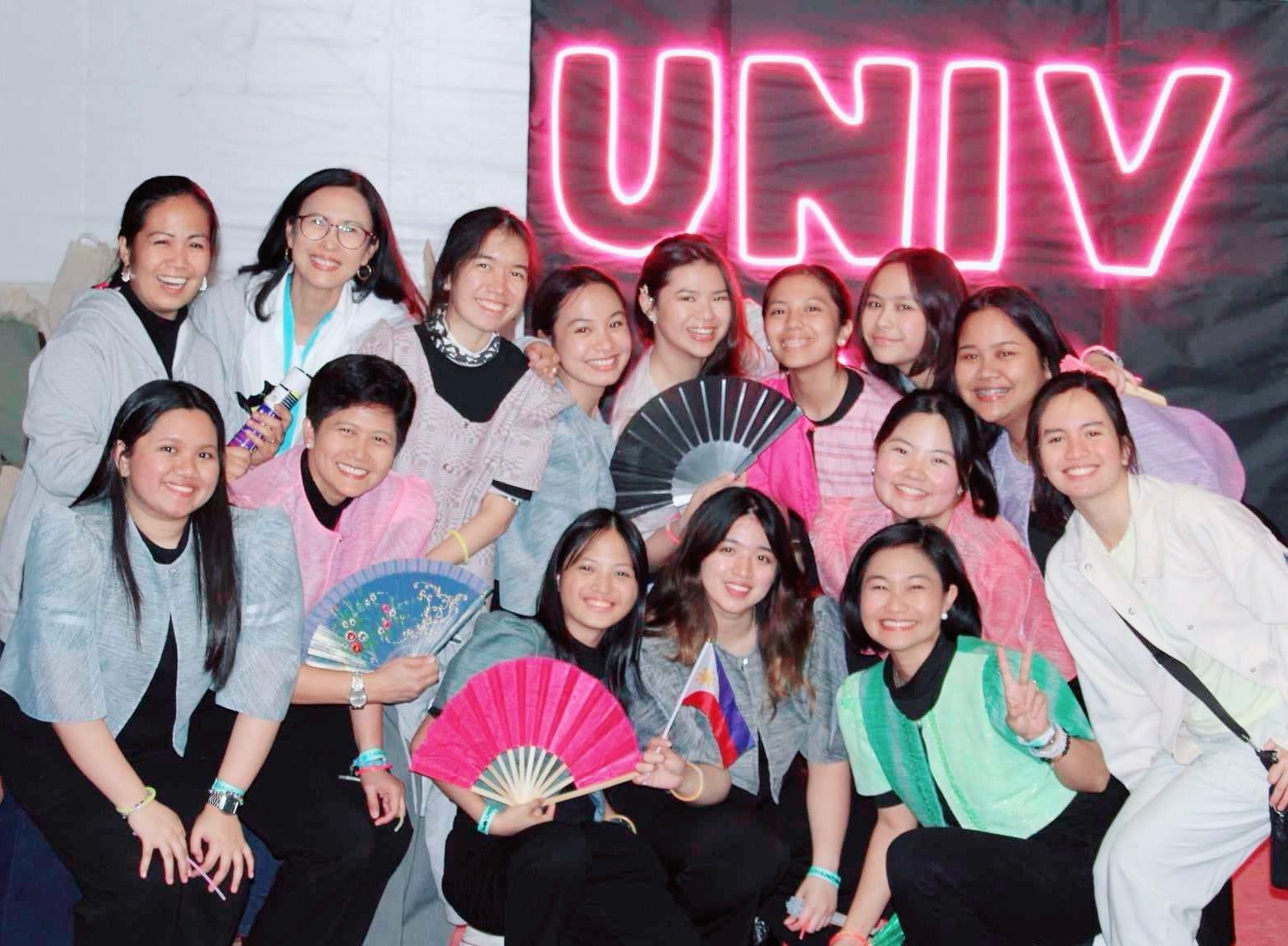 Group of smiling people holding fans in front of a neon UNIV sign.